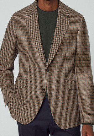 Mann trägt braunen, schwarzen und orange karierten Blazer über einem dunkelgrünen Hemd und marineblaue Hose, Hand in der Tasche.