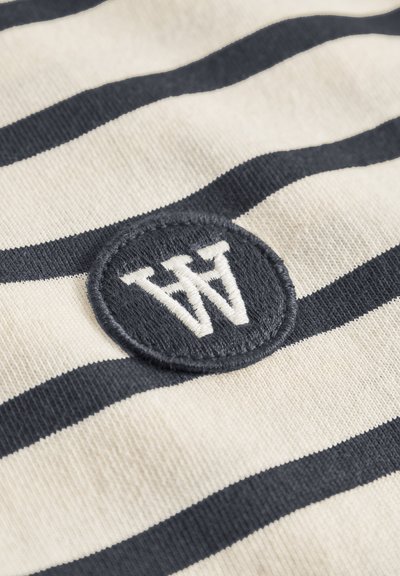 Tissu blanc à rayures horizontales noires avec un patch rond noir brodé de lettres blanches "W" et "M" chevauchantes.