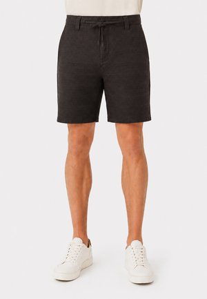 Person trägt schwarze strukturierte Shorts mit Kordelzug in der Taille, ein weißes Hemd, das hineingesteckt ist, und weiße Schnürsneaker und steht vor einem schlichten Hintergrund.