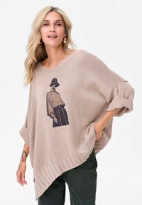 Beżowy sweter z dzianiny o asymetrycznym dolnym wykończeniu, z grafiką postaci w paskowanej bluzce. Żebrowane mankiety i dolny chwyt dodają tekstury.