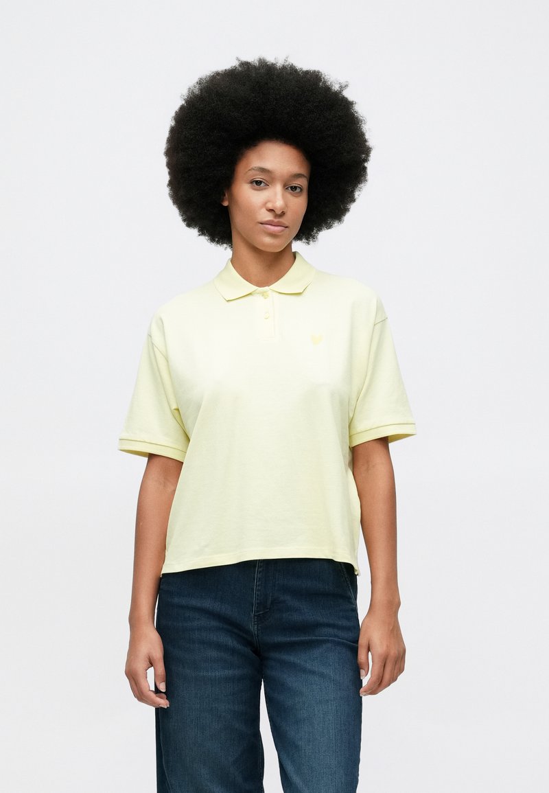 Γυναίκα με αφρο κούρεμα, φορώντας ανοιχτό κίτρινο πόλο μπλουζάκι και σκούρα μπλε τζιν, που στέκεται μπροστά σε λευκό φόντο.