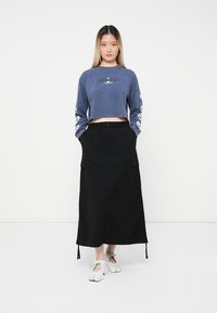 Ženska oblečena v kratek modri dolg rokav top, visok pas črne cargo krila in bele čevlje z razdeljenim prstom, stoji ob belem ozadju.