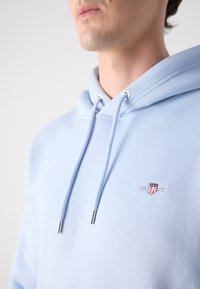 Homme portant un sweat à capuche bleu clair avec cordons et petit logo brodé sur la poitrine, fond blanc.