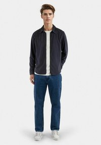 Giacca a zip blu scuro, t-shirt bianca sotto, jeans blu e sneakers bianche; posa in piedi contro uno sfondo neutro.