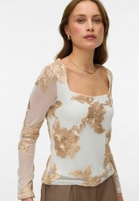 Žena v bílém průsvitném topu s dlouhým rukávem zdobeném béžovou květinovou výšivkou a hnědých kalhotách, doplněná zlatými náušnicemi a náhrdelníkem.