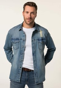 Camisa de mezclilla en azul deslavado, con dos bolsillos en el pecho, cierre de botones en la parte delantera y detalles desgastados en el hombro y la manga.