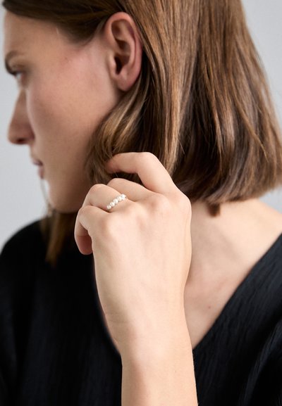 Bague en or ornée de plusieurs petites perles blanches, portée sur une main à la peau naturelle. L'arrière-plan présente un haut texturé en noir.