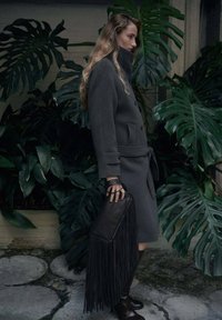 Cappotto grigio in lana con vita con cintura, abbinato a una borsa nera con frange. Modella in piedi vicino a grandi foglie verdi, indossa stivali marroni al ginocchio.