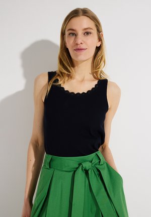 Frau mit langen blonden Haaren, die ein schwarzes ärmelloses Oberteil mit Spitzenbesatz und einen hoch taillierten grünen Rock mit gebundenem Gürtel trägt, steht vor einer weißen Wand.