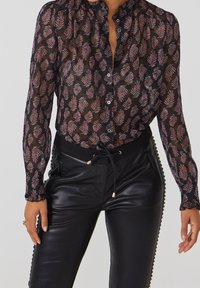 Blusa negra transparente con un patrón floral en púrpura y rosa, que presenta un cuello con volantes y mangas largas, combinada con pantalones negros.