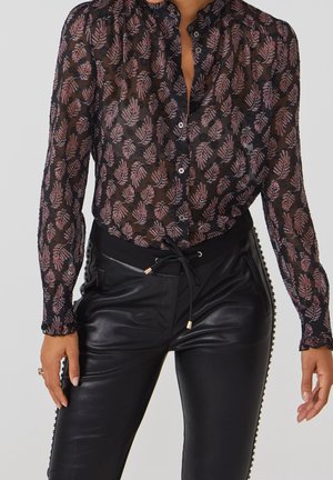 Blouse noire transparente avec un motif floral en violet et rose, dotée d'un col froncé et de longues manches, assortie à un pantalon noir.