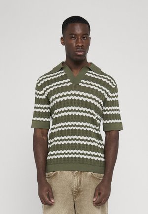 Mann trägt olivgrün-weiß gestreiftes gestricktes Poloshirt mit kurzen Ärmeln und beiger Hose, steht vor hellem einfarbigem Hintergrund.