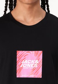 Musta t-paita, jossa on neliömäinen grafiikka vaaleanpunaisella taustalla ja valkoisella tekstillä "JACK & JONES CORE UNLTD." Pehmeää kangasta, pyöreä pääntie.