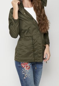 Kobieta w oliwkowej zielonej kurtce z kapturem, z kieszeniami z przodu, oraz niebieskich dżinsach z czerwonymi haftami kwiatowymi na jednej nogawce.