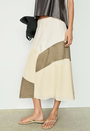 Frau trägt einen Midi-Rock in Beige und Braun mit Farbblock, ein braunes ärmelloses Oberteil, hält eine schwarze geflochtene Tasche und trägt beige flache Sandalen.