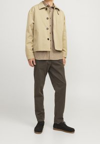 Homme portant une veste beige boutonnée sur une chemise à carreaux, un pantalon marron foncé et des chaussures noires, debout devant un fond clair uni.