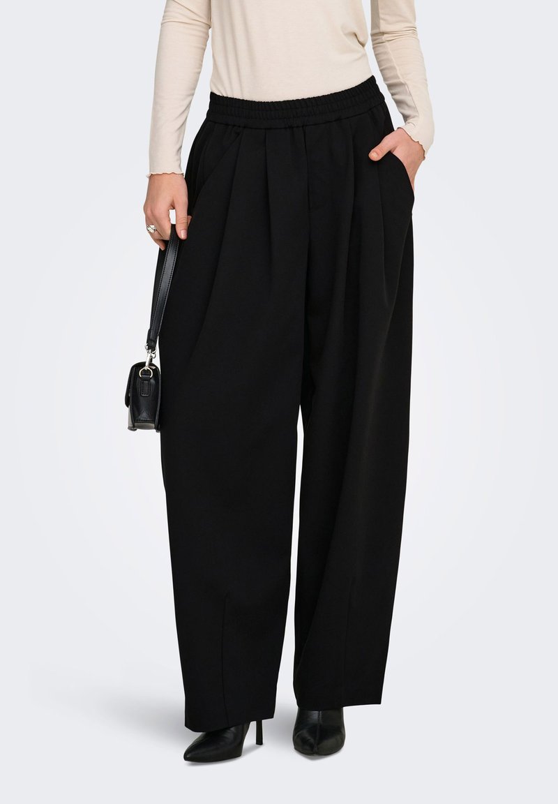 Γυναίκα που φοράει μαύρο παντελόνι ψηλόμεσο, με φαρδιά μπατζάκια και ελαστική μέση, μπεζ μακρυμάνικη μπλούζα, μαύρα μποτάκια με τακούνι, κρατώντας μια μικρή μαύρη τσάντα.