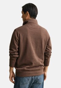 Maglione marrone con collo alto e polsini a coste, realizzato in materiale morbido. La parte posteriore presenta una texture liscia e dettagli di cuciture sottili.