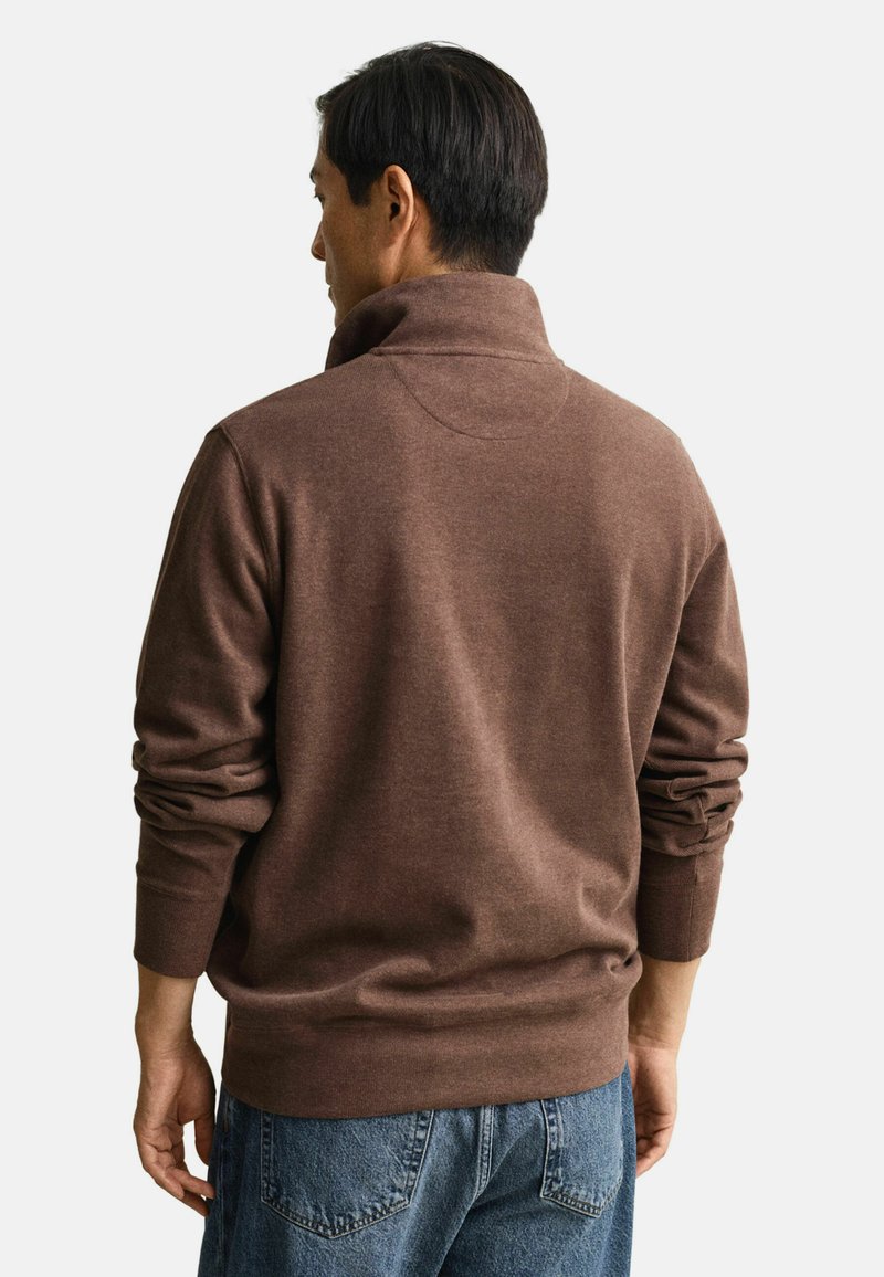 Brun tröja med hög krage och ribbade ärmslut, gjord av mjukt material. Baksidan har en slät textur och subtila sömdetaljer.