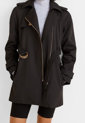 Kvinde iført en sort trenchcoat med bælte og guld lynlåse over en hvid rullekrave og sorte knælange shorts.