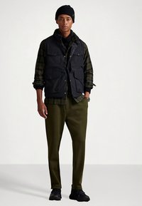 Melnā veste ar vairākām kabatām pār tumši zaļu pledveida kreklu, olīvu krāsas bikses un melni sporta apavi, aksesuāros melna cepure.