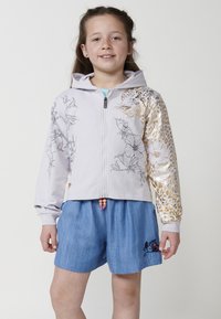 Svijetlosiva kapuljača s floralnim i zlatnim printom leopard uzorka, koja ima rajsferšlus i kapuljaču. Uparena s plavim traper kratkim hlačama s vezom.