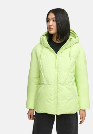 Vrouw in een lichgroene pufferjack met capuchon, dichtgeritst, staand met ontspannen handen langs haar zij, kijkend naar rechts.