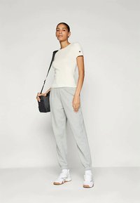 Creme tilpasset t-shirt, grå sweatpants og hvide sneakers. Modellen holder en lille sort taske. Simpelt design uden mønstre eller udsmykninger.