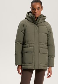 Olive zöld puffermellény kapucnival, vízszintes steppelésű kialakítással, két elülső zsebből és cipzáras záródással. Puha tapintás.