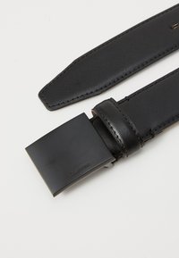 Cinturón de cuero negro con una hebilla cuadrada de color negro mate. Costura visible a lo largo de la correa, con un logo de Calvin Klein en la hebilla.
