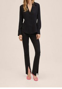 Blouse noire à manches longues avec patte de boutonnage, associée à un pantalon noir ajusté avec fentes latérales, complétée par des talons à brides roses.