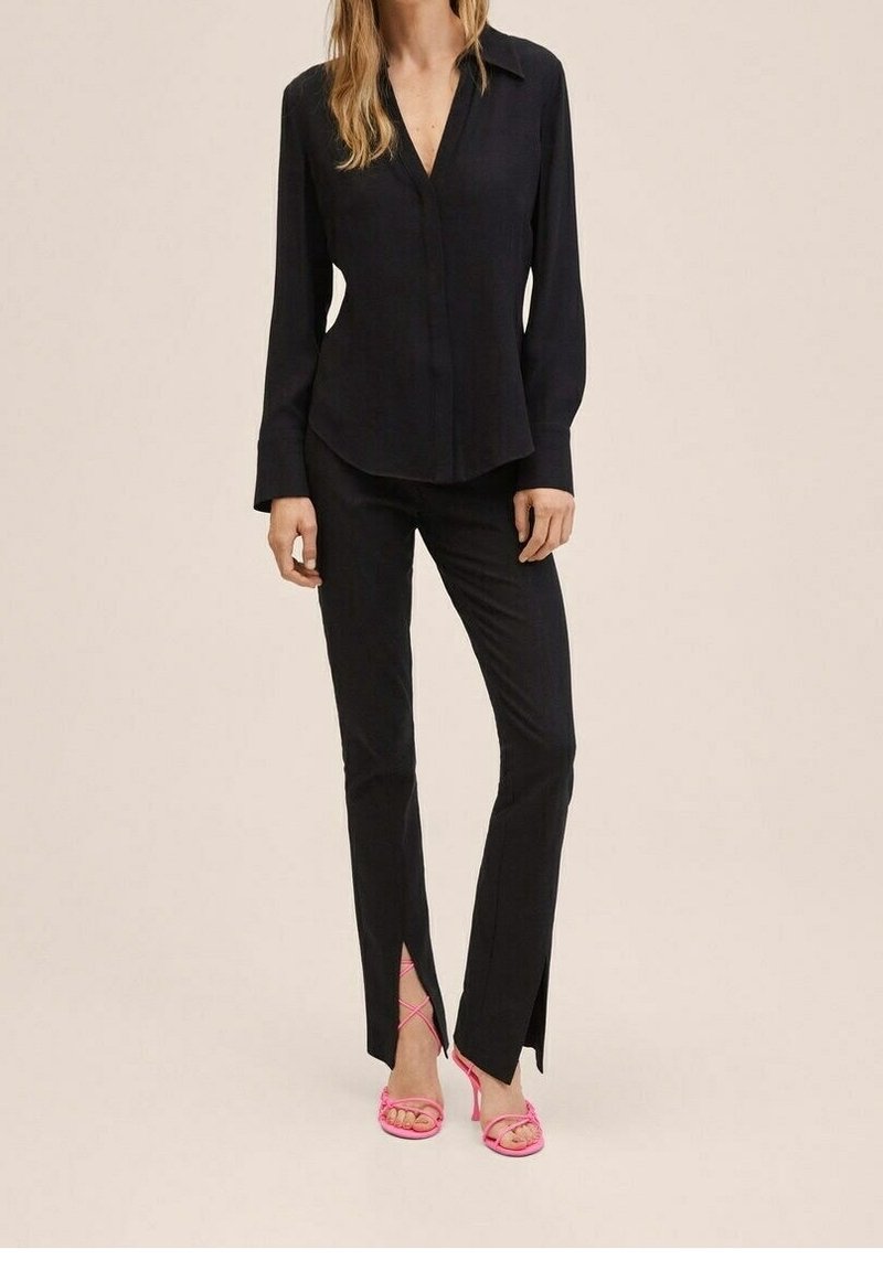 Blouse noire à manches longues avec patte de boutonnage, associée à un pantalon noir ajusté avec fentes latérales, complétée par des talons à brides roses.
