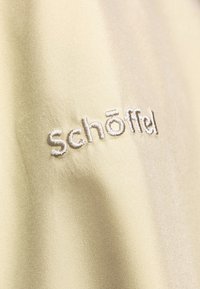 Heller beige Stoff mit glatter Textur zeigt das gestickte "schöffel"-Logo in silbernem Faden, das auffällig auf der Oberfläche platziert ist.