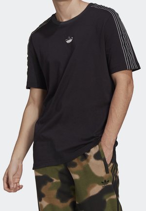 Mężczyzna ubrany w czarną koszulkę Adidas z białymi paskami na rękawach oraz krótkie spodenki Adidas w kamuflażowy wzór, z ręką w kieszeni.