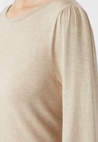 Beige svetr s kulatým výstřihem a jemně nařasenými rukávy. Textura vypadá hladce a pleteně, s jemnými variacemi v barvě.