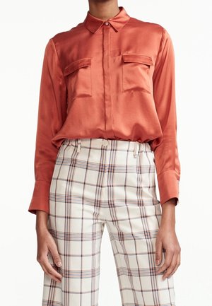 Blouse en satin couleur rouille à manches longues avec poches poitrine boutonnées, associée à un pantalon blanc à carreaux taille haute.