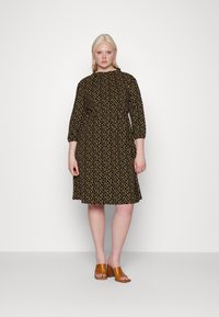 Vestido preto com bolinhas amarelas, decote franzido e mangas três quartos. O modelo usa sandálias abertas castanhas. Tecido suave, caimento descontraído.