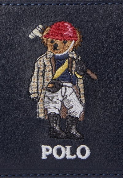 Medvedík v polohovom oblečení s červenou prilbou, károvanou bundou, bielymi nohavicami, topánkami a držiaci palicu na polo, s textom „POLO“ pod ním.