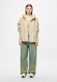 Beežikarva voodriga jope, kus on tikitud paneelid, kombineeritud helepruuni argyle-villase kampsuni ja lahtiste roheliste teksapükstega. Omab kuldse luku tõmbenuppu.