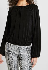 Czarna bluzka z długim rękawem, z marszczonym dekoltem i zwężanym dołem, w połączeniu z szarymi spodniami w wężowy wzór o gładkiej teksturze.
