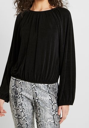 Blouse noire à manches longues avec un col froncé et un ourlet cintré, associée à un pantalon gris à imprimé serpent au tissu lisse.