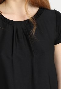 Kobieta ubrana w czarną bluzkę z krótkim rękawem z marszczonym dekoltem i luźnym krojem, odsłaniającą górną część klatki piersiowej i częściowo ramię.