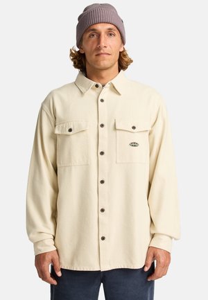Long-sleeve, cream-colored shirt made of soft fabric with two chest pockets and brown buttons; worn with a light purple knit beanie.