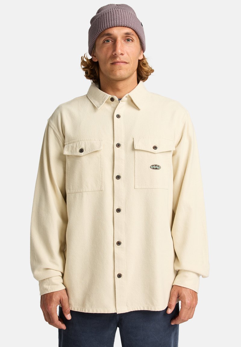 Lange mouwen, crèmekleurig shirt van zachte stof met twee borstzakken en bruine knopen; gedragen met een lichtpaarse gebreide beanie.