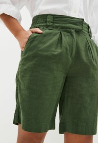 Grüne Leinen-Shorts mit Falten, elastischem Bund, Seitentaschen und rohem Saum. Die Stoffstruktur wirkt leicht und atmungsaktiv.