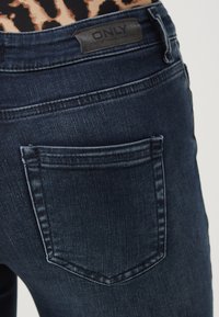 Un bolsillo de jeans de mezclilla azul oscuro sobre una persona que lleva una camiseta de leopardo, con una etiqueta de marca de cuero negro "ONLY" visible en la cinturilla.