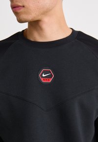Sort sweatshirt med en hexagonal Nike-logo patch i rød og hvid. Har en tekstureret, glat overflade og raglanærmer.