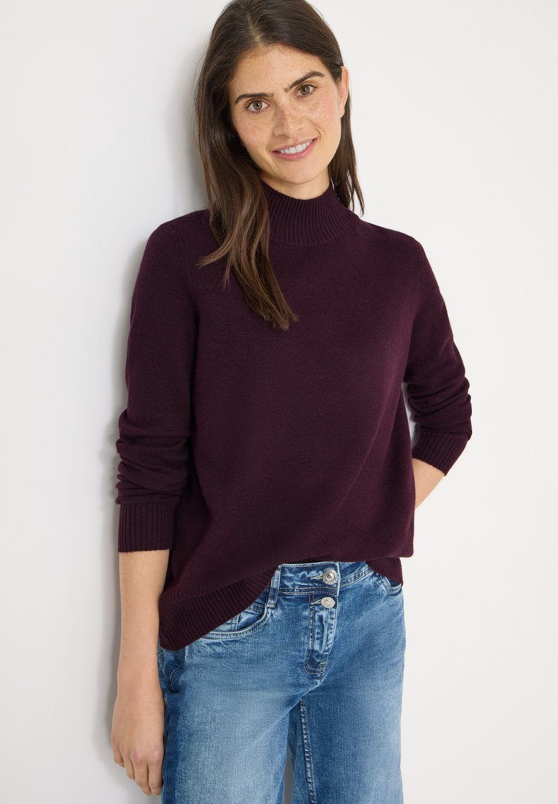 Bordeauxroter Pullover mit hohem Kragen und gerippten Bündchen, kombiniert mit hellblauen Jeans mit einem ausgewaschenen Finish und doppelter Knopfverschluss.