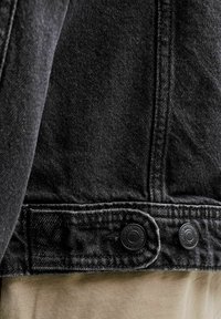 Nærbilde av hem på svart denimjakke med to svarte metallknapper og synlige sømmer over beige stoff.