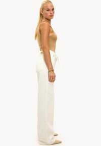 Modelo vistiendo una camiseta sin mangas ajustada beige y unos pantalones de mezclilla blancos de pierna ancha de cintura alta con un cinturón atado, complementados con pulseras y anillos de oro.